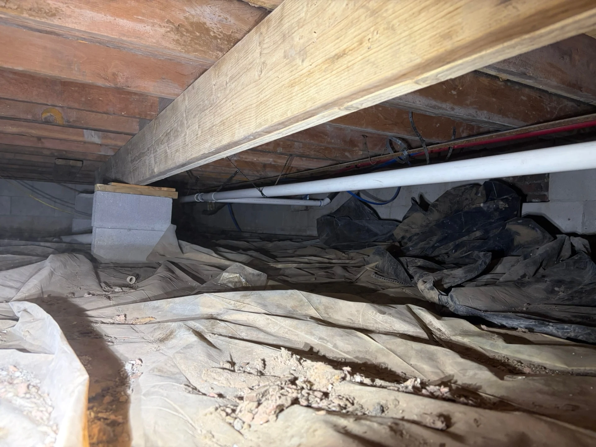 Crawlspace drain repipe showing PVC drain line and PEX water lines secured to wooden floor joists in Rogers, Arkansas.